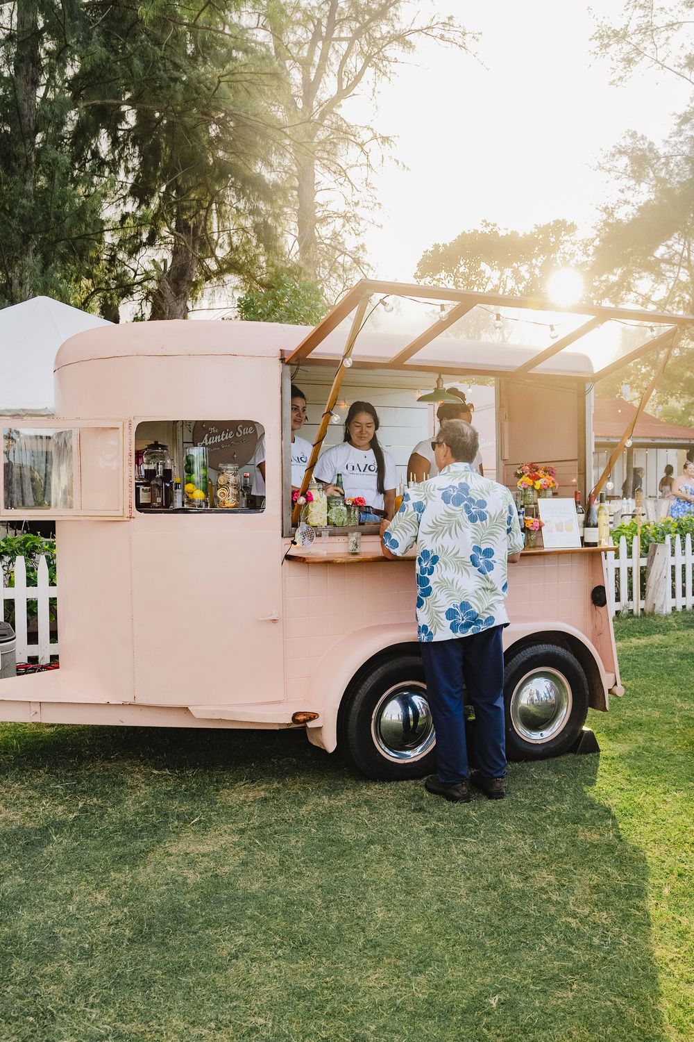 A pink vintage food truck serving customers at an outdoor event during golden hour sunset.