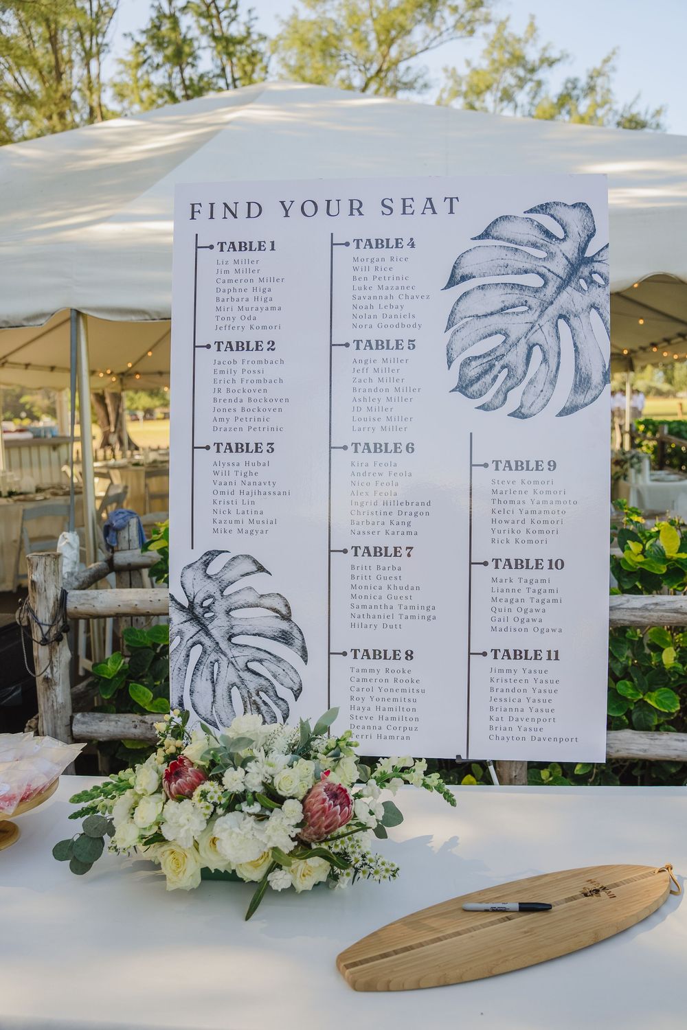 Wedding seating chart with tropical monstera leaf design displayed on surfboard with elegant floral arrangements.