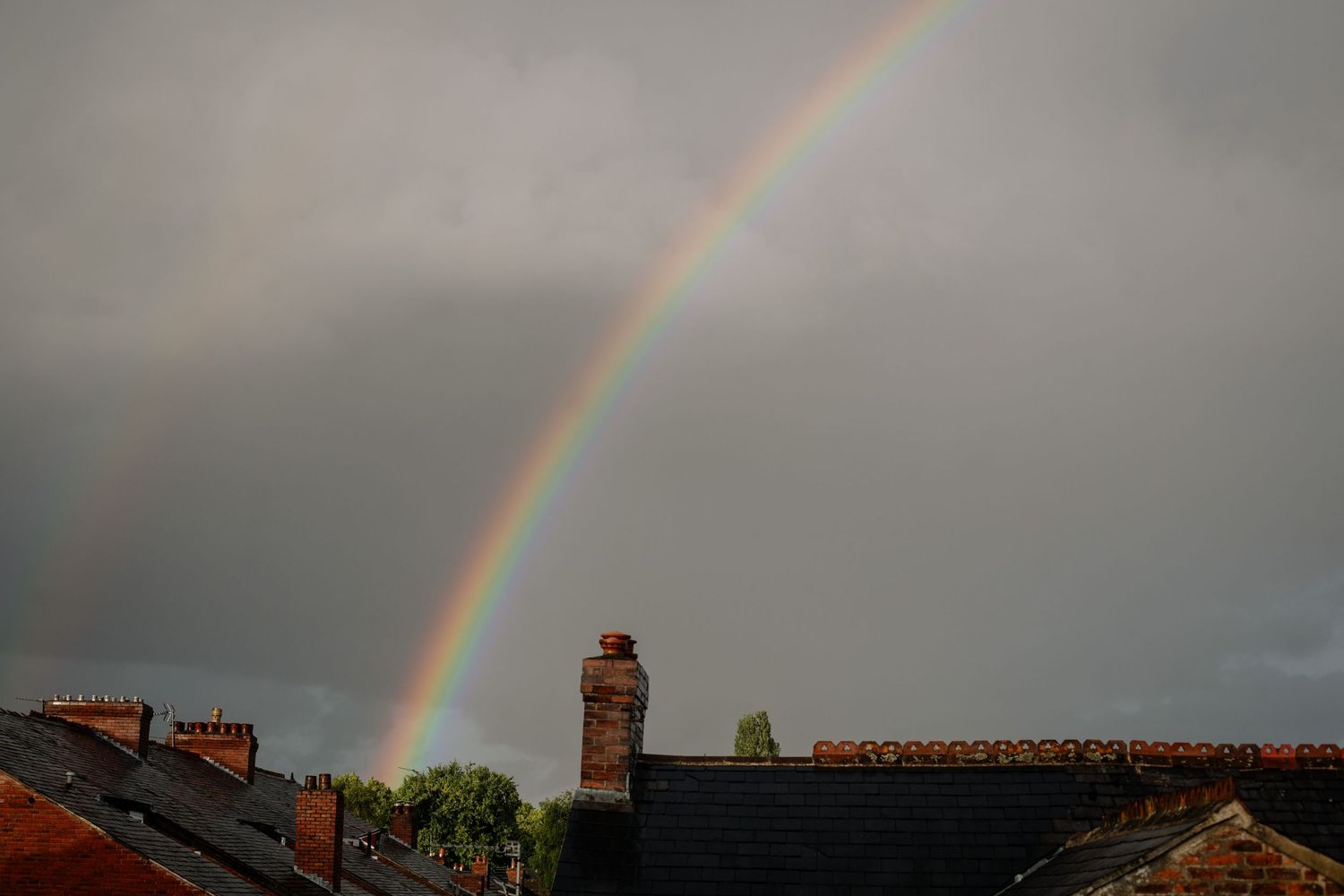 Vibrant rainbow arches over residential rooftops against a moody grey sky.