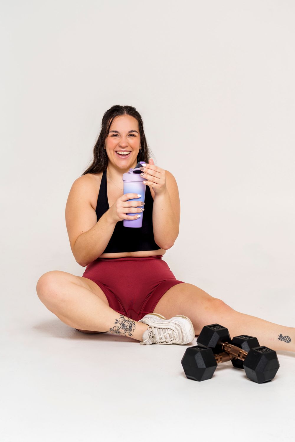 Woman in workout attire relaxing with protein shake and dumbbells after exercise session.