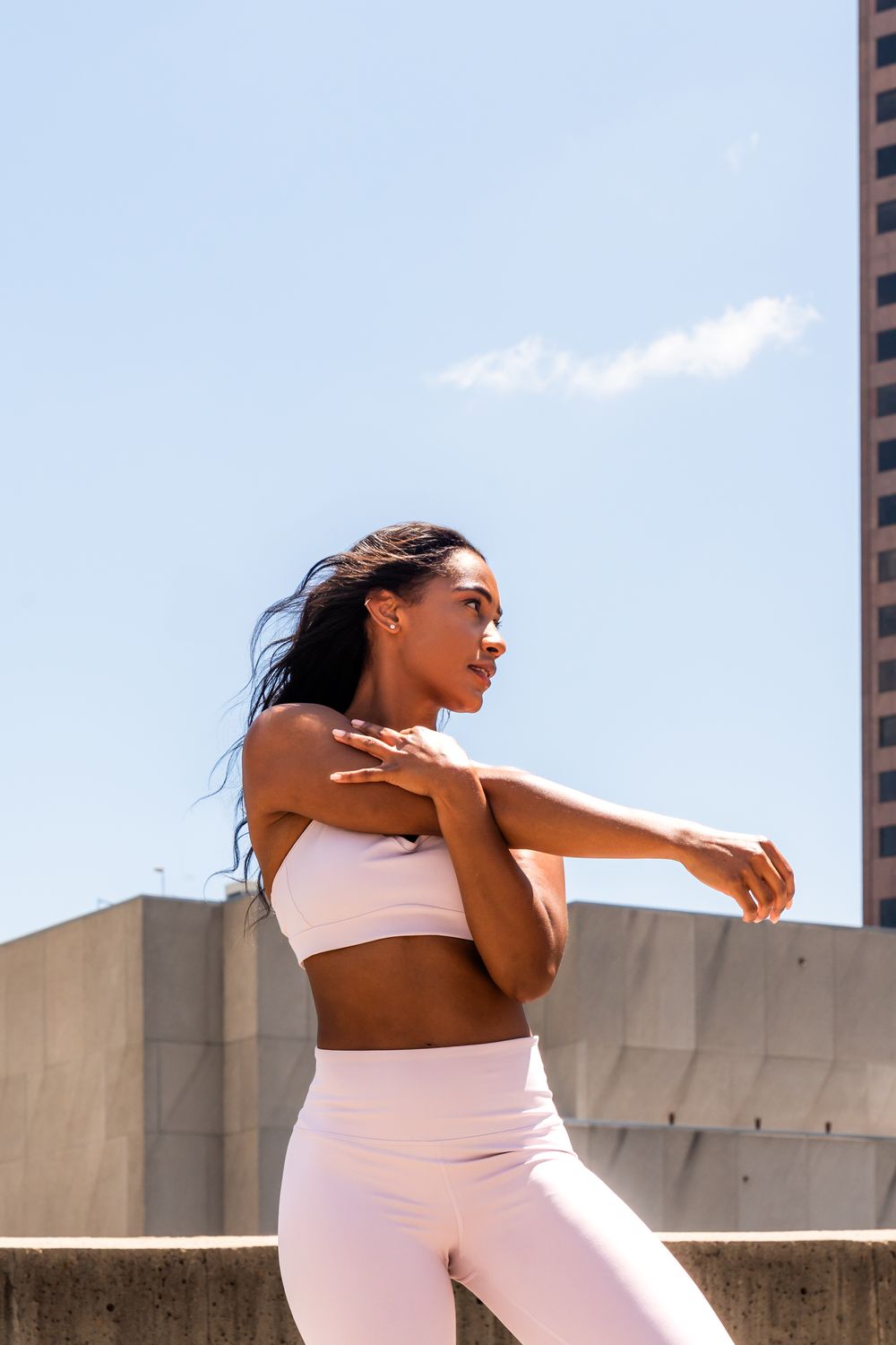 Athletic woman in pink workout set stretching outdoors against urban backdrop.