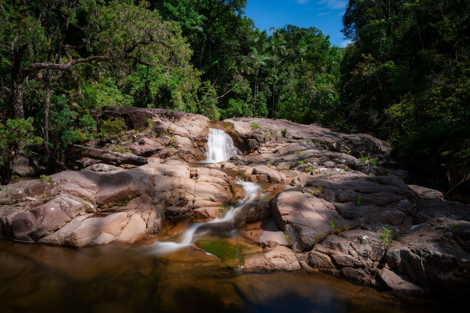 Crystal clear waterfall cascades over sun-lit rocks in a tropical forest setting.
