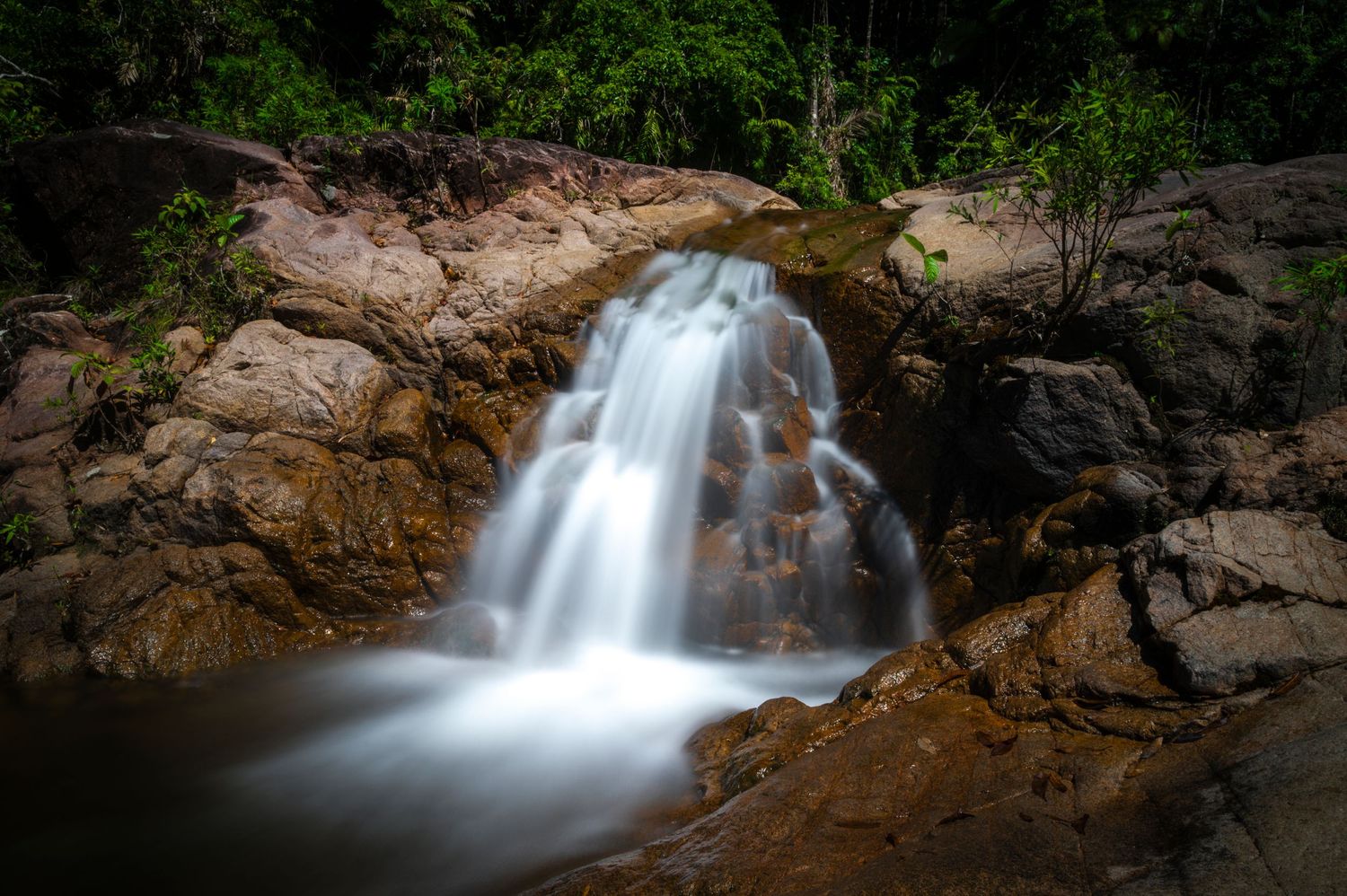Silky waterfall cascades over golden rocks surrounded by dense tropical vegetation.