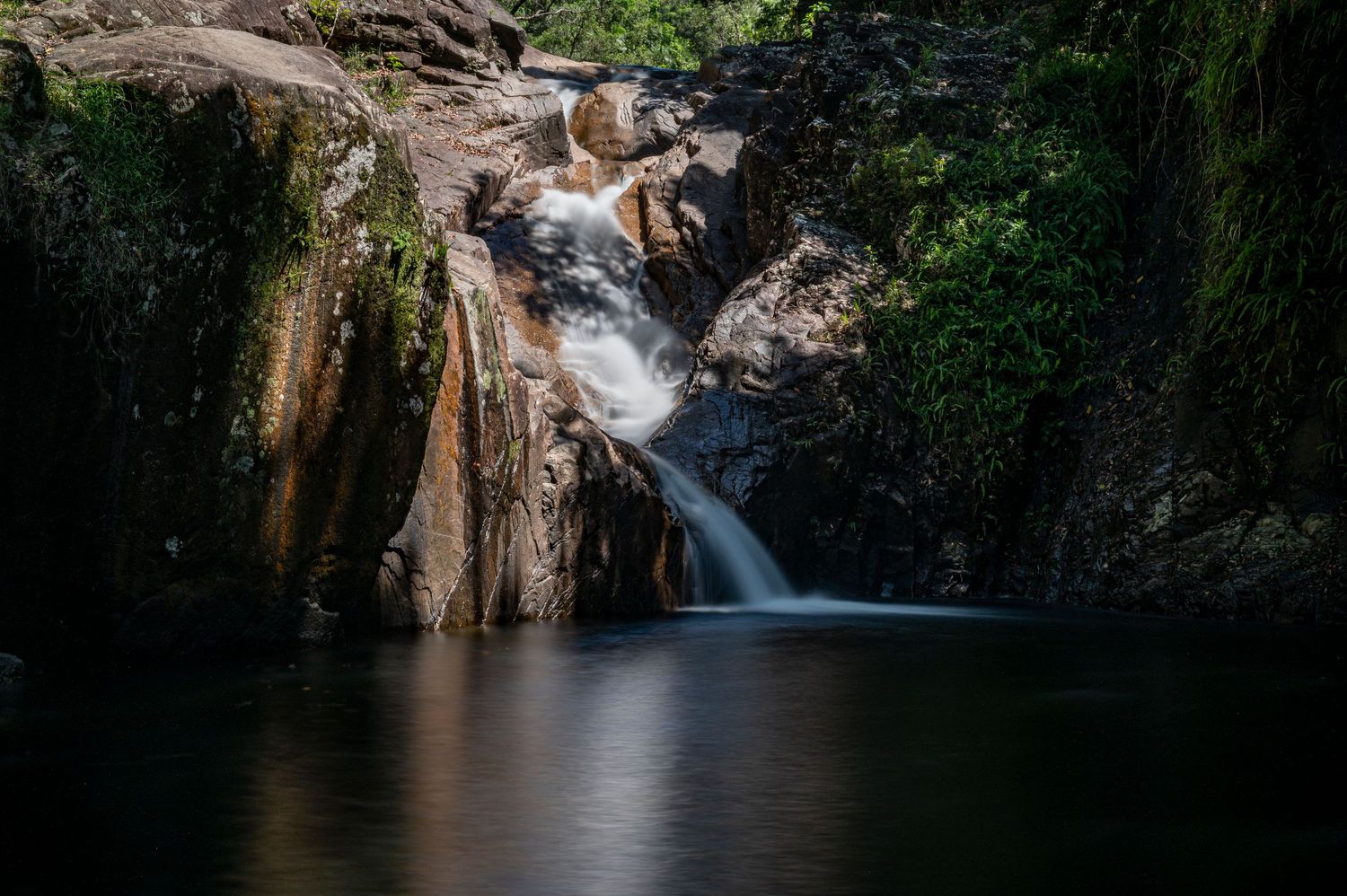 Long exposure waterfall flows smoothly through dark rocky cliffs in dense forest.