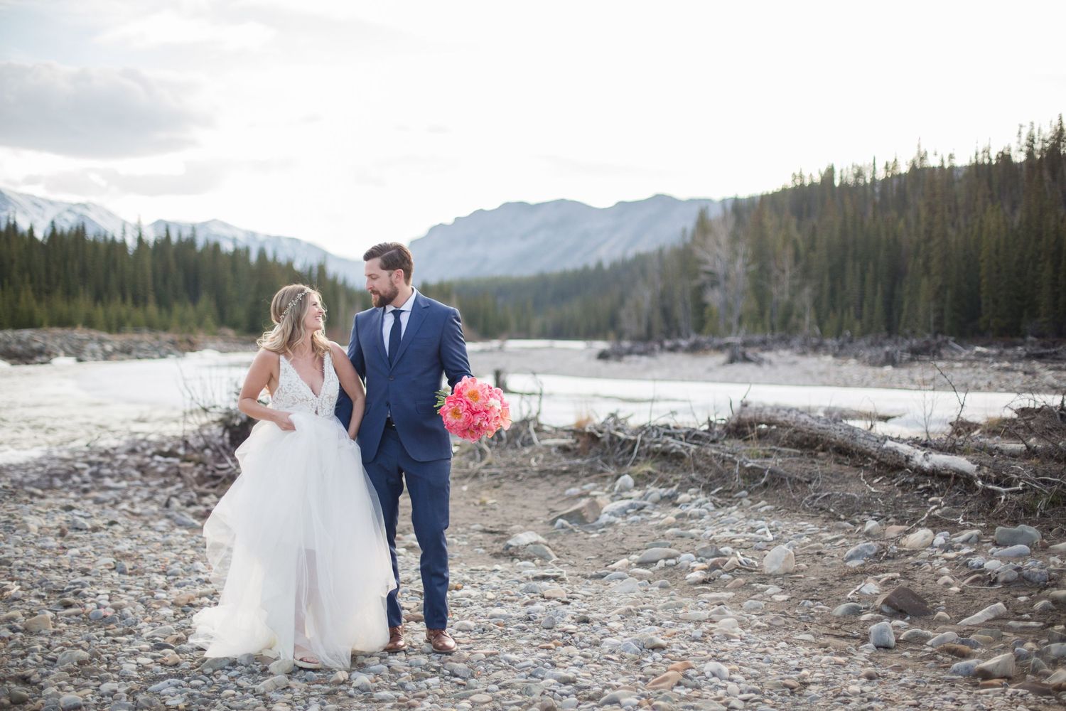 Wedding couple walking together on rocky riverbed with mountain landscape in background.