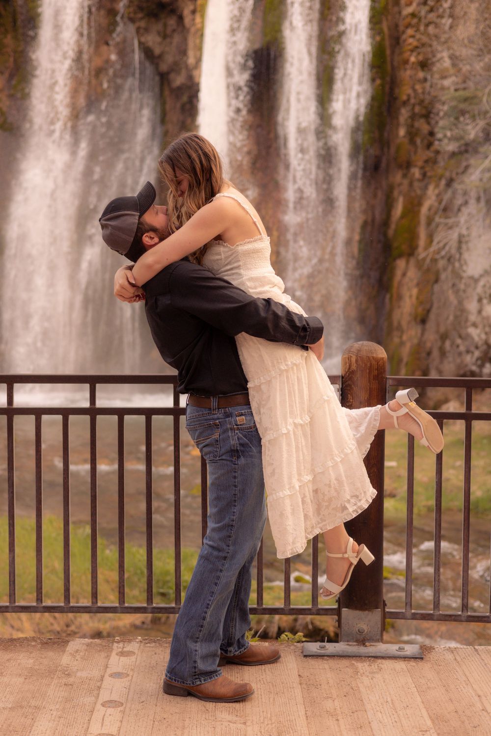 A romantic couple embraces in front of a majestic waterfall while standing on a wooden viewing platform with railings.