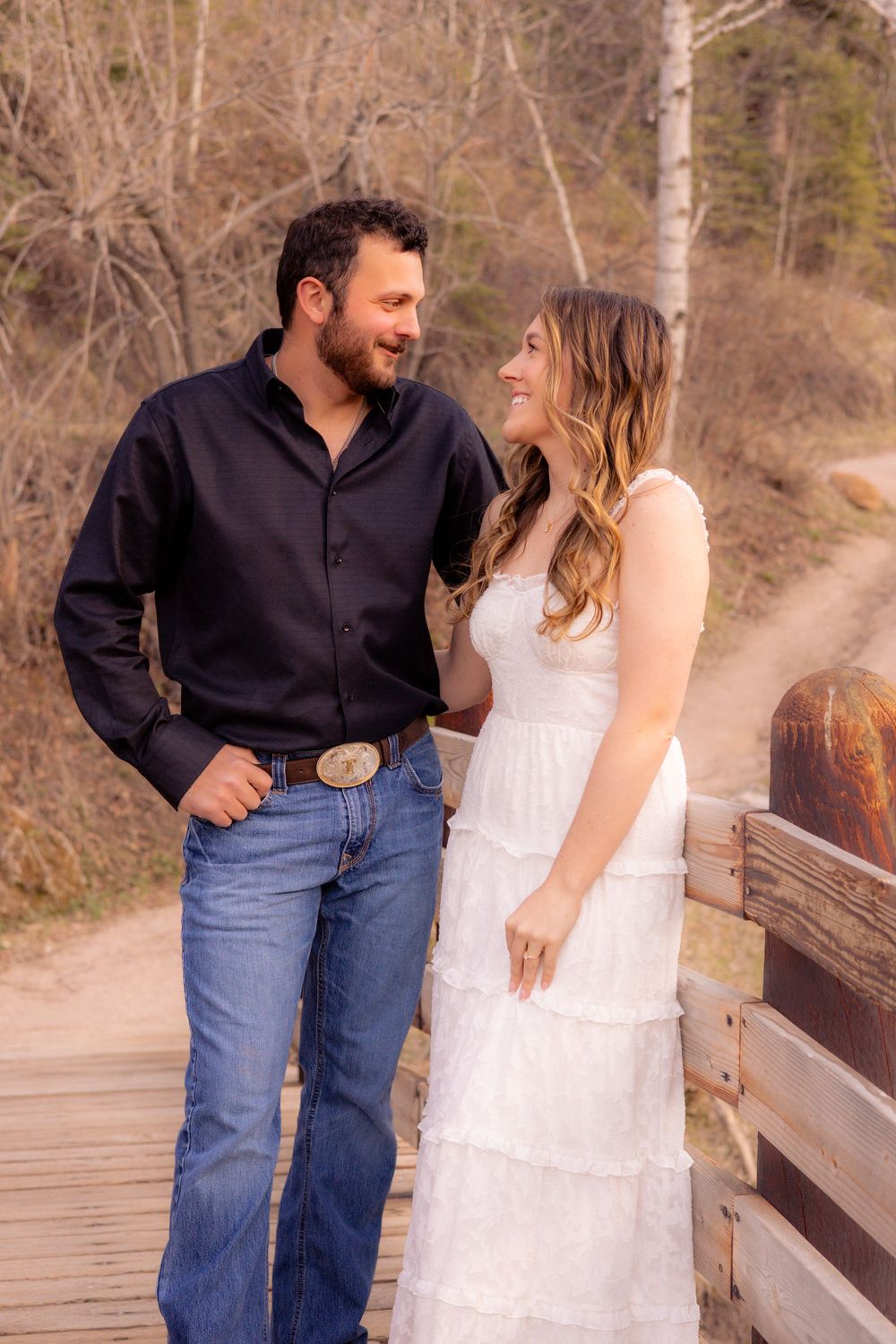 A couple stands together on a wooden walkway, with the woman in a white dress and the man in a dark shirt and jeans.