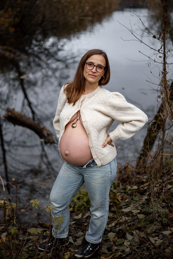 A pregnant person in a cream sweater and jeans poses by a lake on an autumn day.
