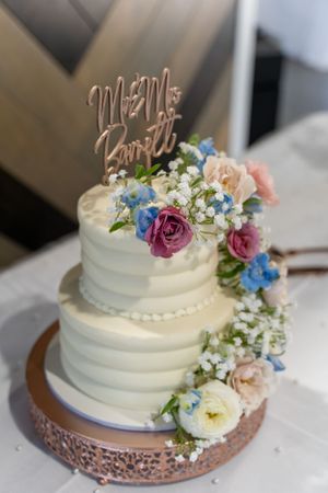 Two-tiered white wedding cake decorated with fresh pink, purple and blue flowers on a rose gold stand.