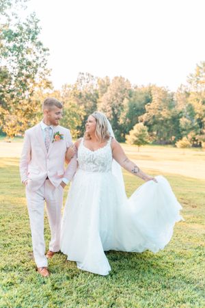 Couple in light-colored wedding attire poses playfully on sunlit grass.
