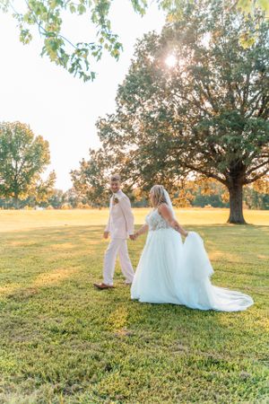 Newlyweds walking hand in hand through a sun-dappled field at golden hour.