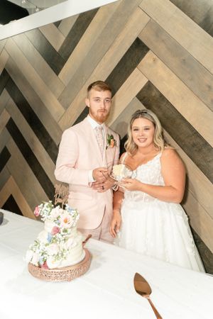 Couple cuts wedding cake in front of geometric wooden wall pattern at reception.