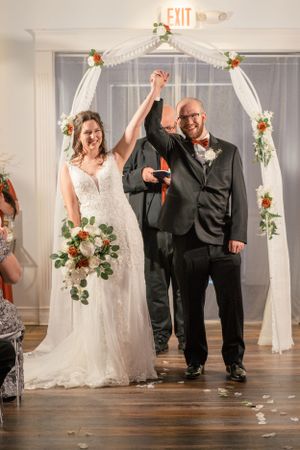 A wedding ceremony under a white floral arch decorated with roses and greenery.
