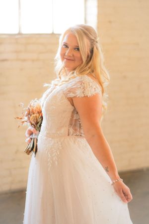 Bride in lace wedding dress poses with bouquet in sunlit indoor setting.