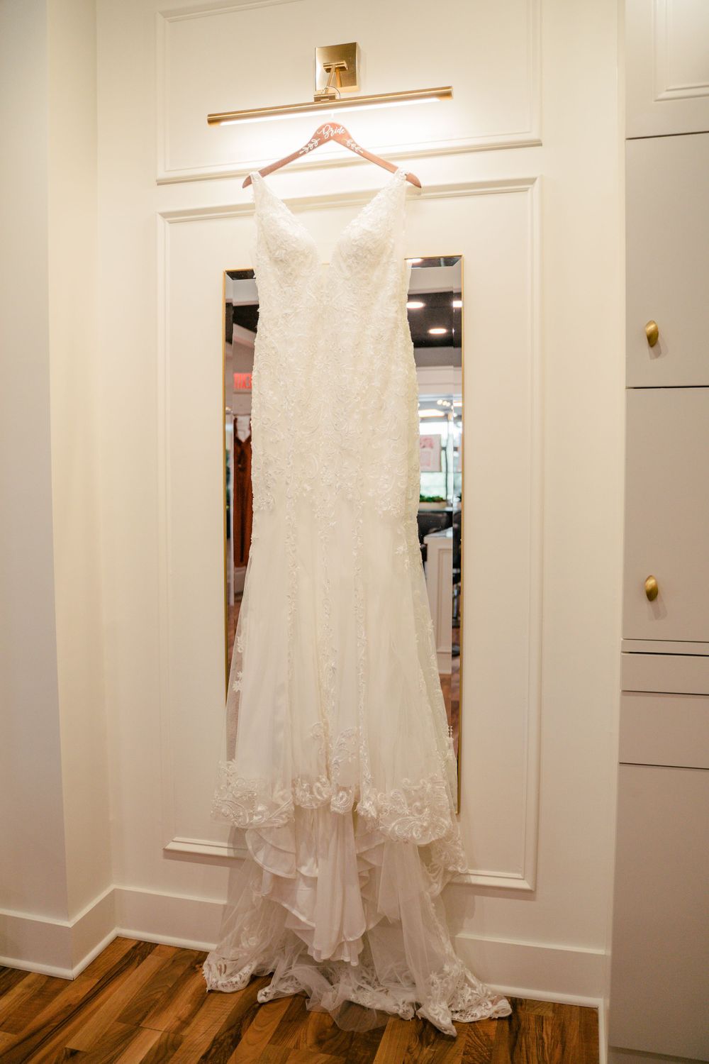An elegant lace wedding dress hanging on display in a white room.