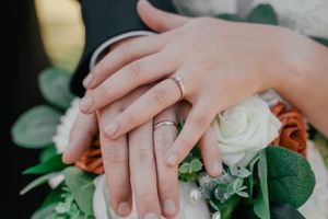Close-up of hands with wedding rings resting on a bouquet with white and peach roses and greenery.