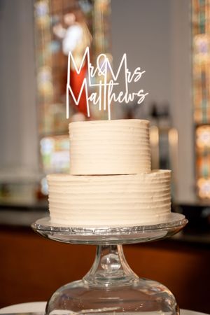 White two-tier wedding cake with 'Mr & Mrs Matthews' custom acrylic topper on glass stand.