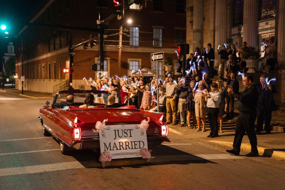 Wedding party riding in red convertible at night with crowd cheering along street.