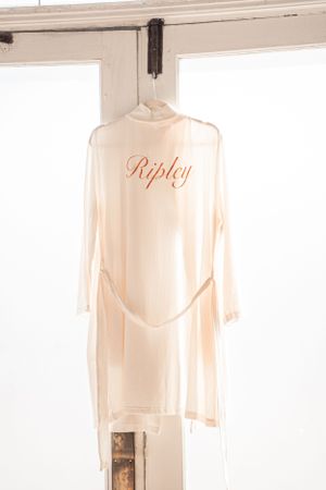 A sheer white bridal robe with 'Ripley' monogrammed in orange hanging by a window.