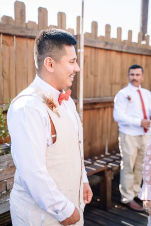 A groom in a light vest and bow tie smiling during an outdoor wedding ceremony.