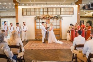 Wedding ceremony in an indoor venue with hanging lights and wooden beams overhead as attendants watch.