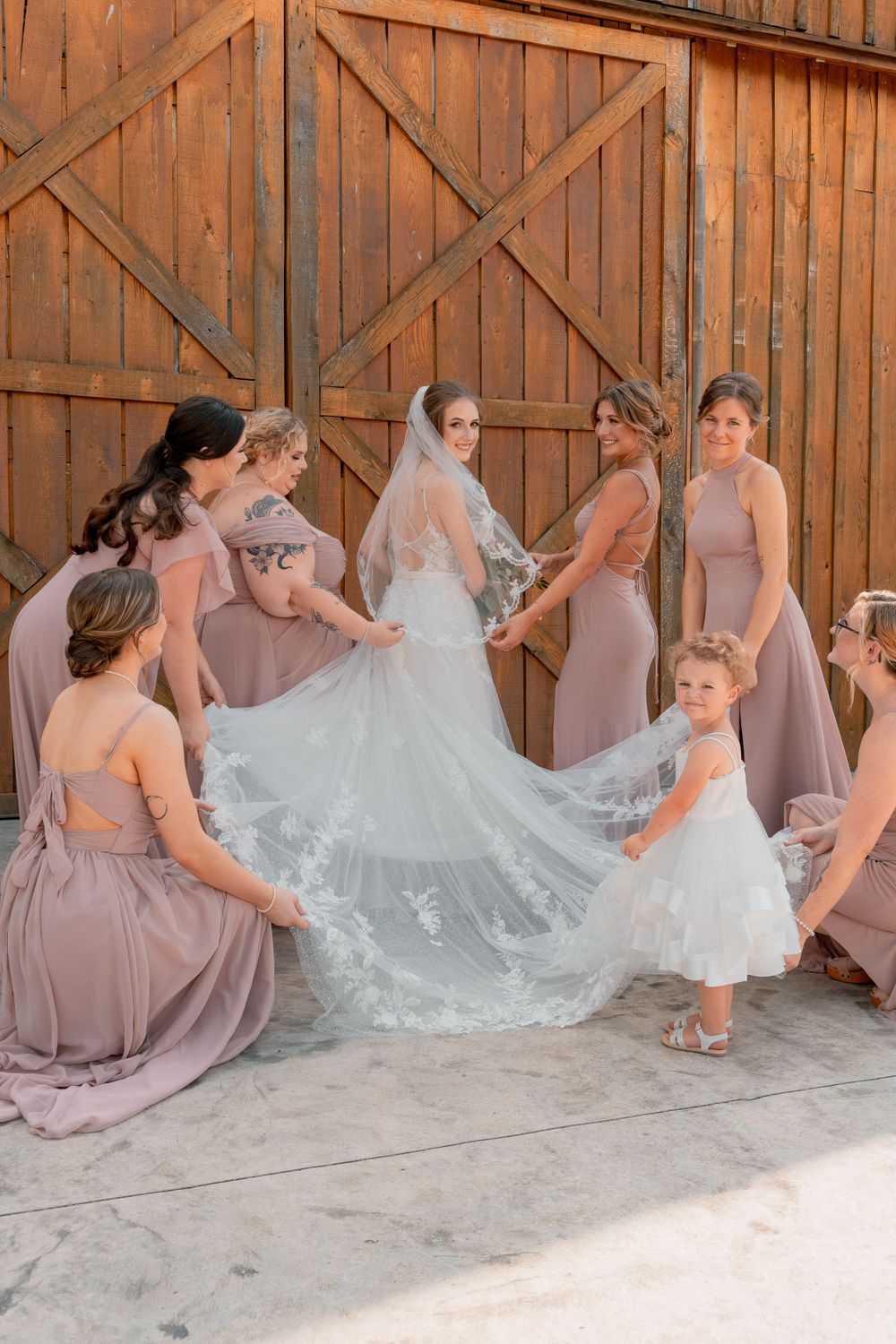 Bridesmaids in dusty rose dresses arrange a bride's lace train outside a rustic barn.