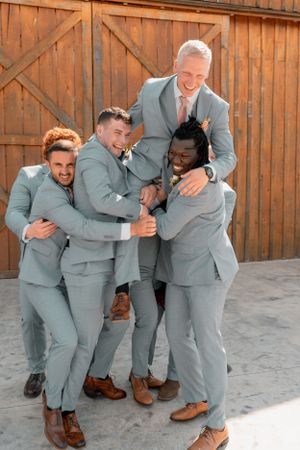 Groomsmen in light gray suits share a playful group hug against wooden barn backdrop.