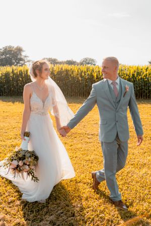 Newlyweds walking hand in hand through golden field at sunset.