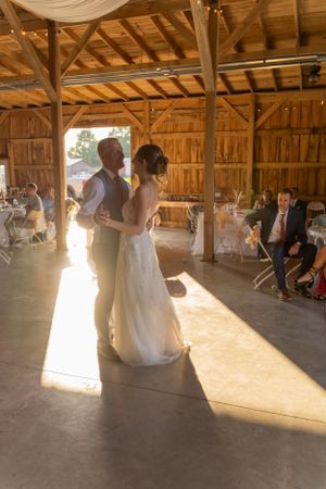 A couple shares their first dance in a rustic barn venue with sunlight streaming through the entrance.