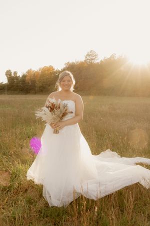 Bride holding dried flower bouquet in sunlit field wearing flowing white gown.