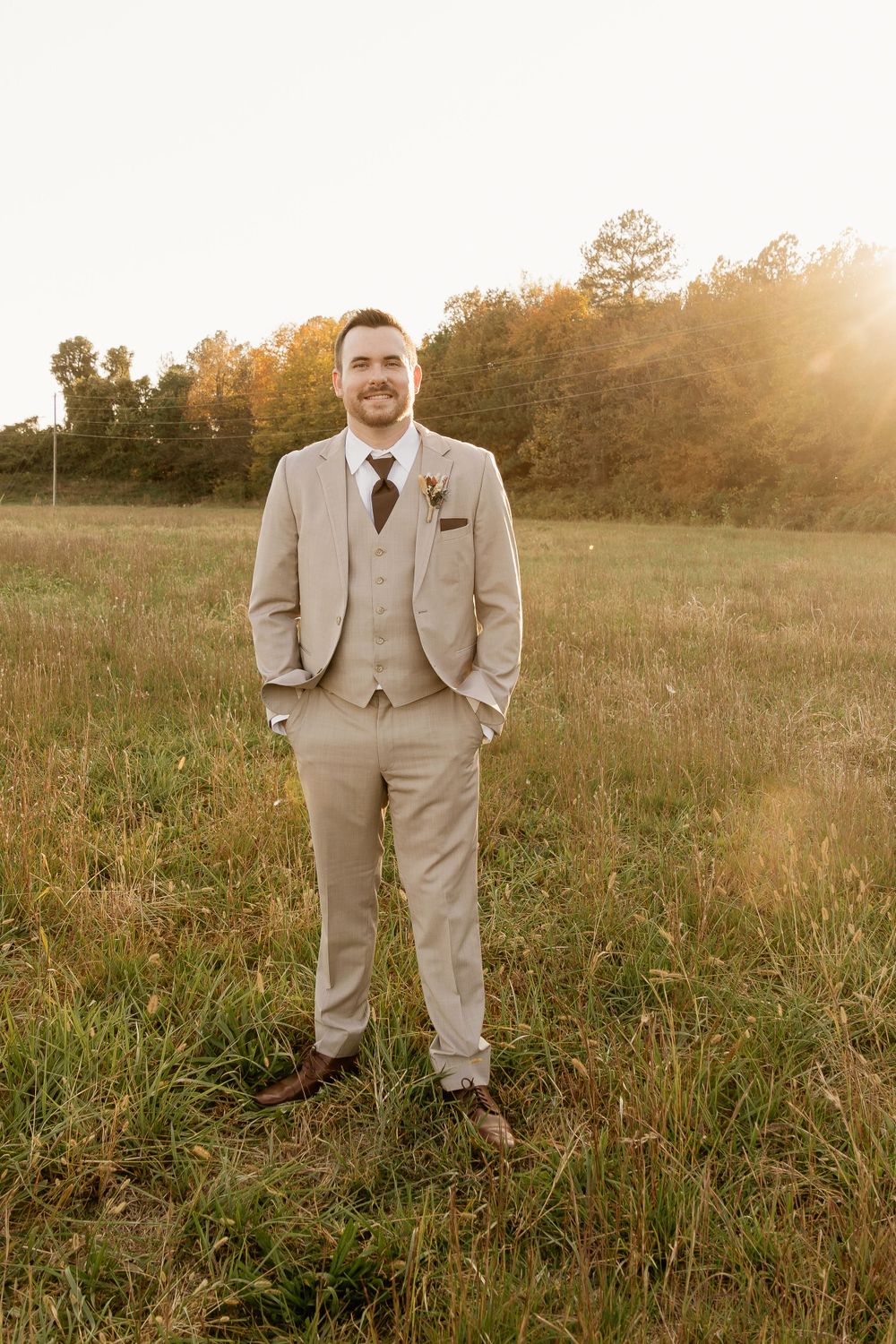 Groom in beige suit standing in golden field during sunset.