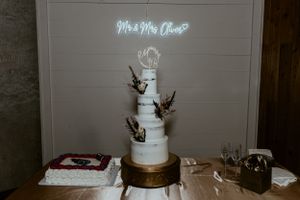 Elegant white wedding cake with branches and neon 'Mr & Mrs' sign displayed on gold stand at reception.