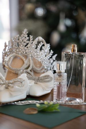 Elegant bridal accessories including a crystal tiara, sparkly white shoes, and perfume bottles arranged on a green surface.