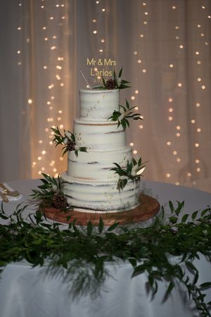 Elegant three-tier white wedding cake decorated with greenery against a backdrop of twinkle lights.