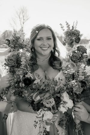 A beaming bride in white holds up two vibrant floral bouquets in an outdoor setting.