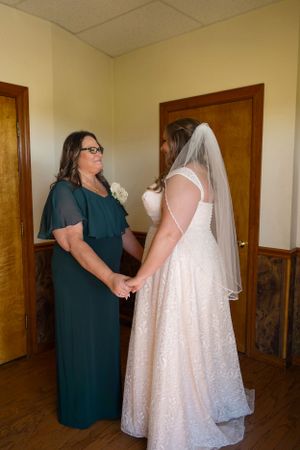 An emotional moment between a bride in white and woman in teal dress in a simple room.