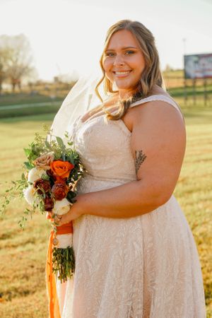 Bride in lace dress holding autumn-colored bouquet during golden hour outdoor portrait.