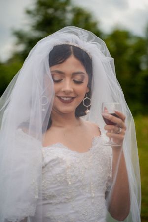 Elegant bride in lace dress and veil holding champagne glass outdoors.