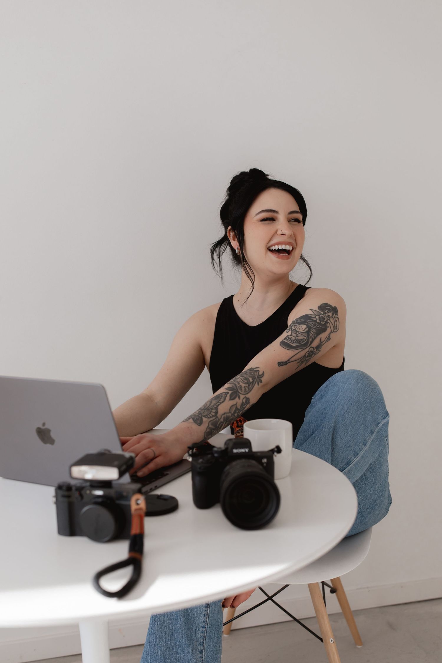 Photographer with tattoos laughs while working on laptop at white desk with cameras and coffee cup.