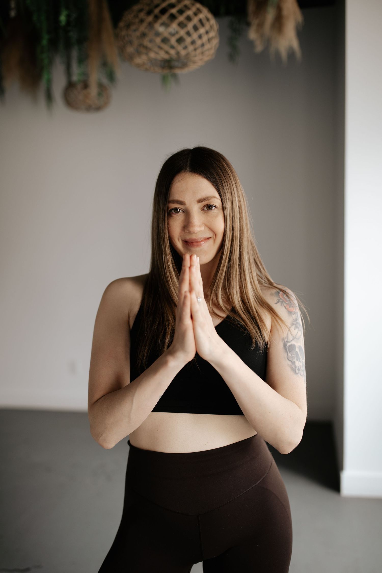 A person in black workout attire stands in prayer pose during yoga practice, with woven baskets hanging in background.