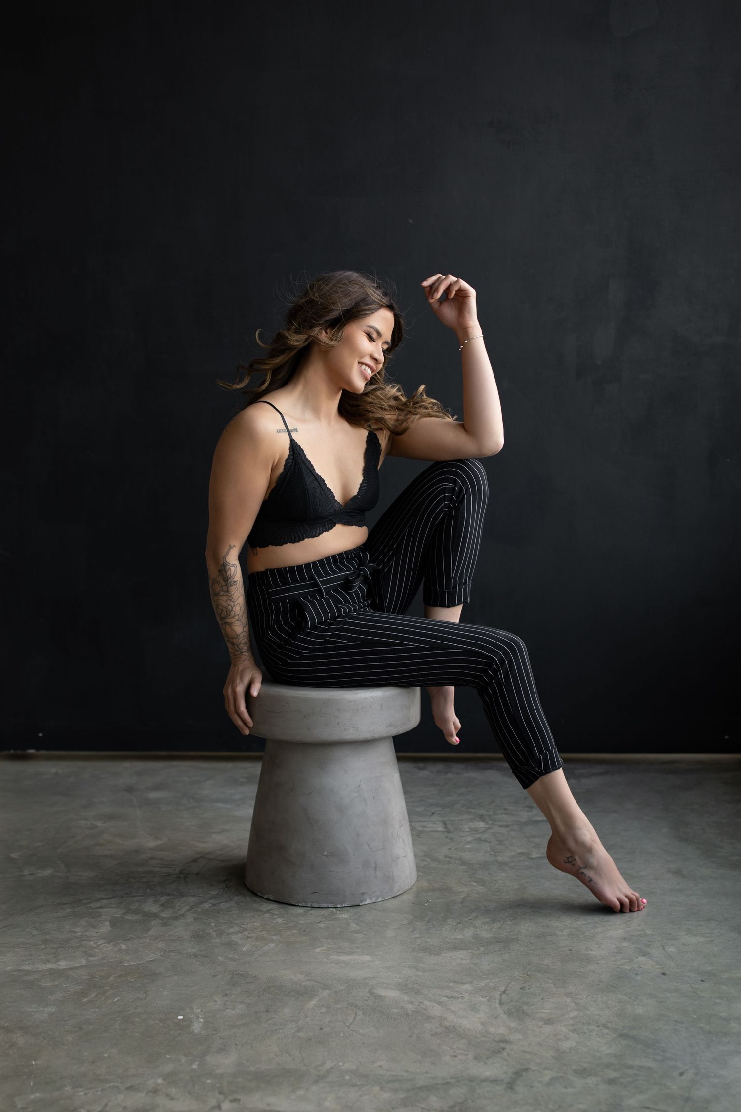 A person in striped pants and black top sits casually on a concrete stool against a dark wall.