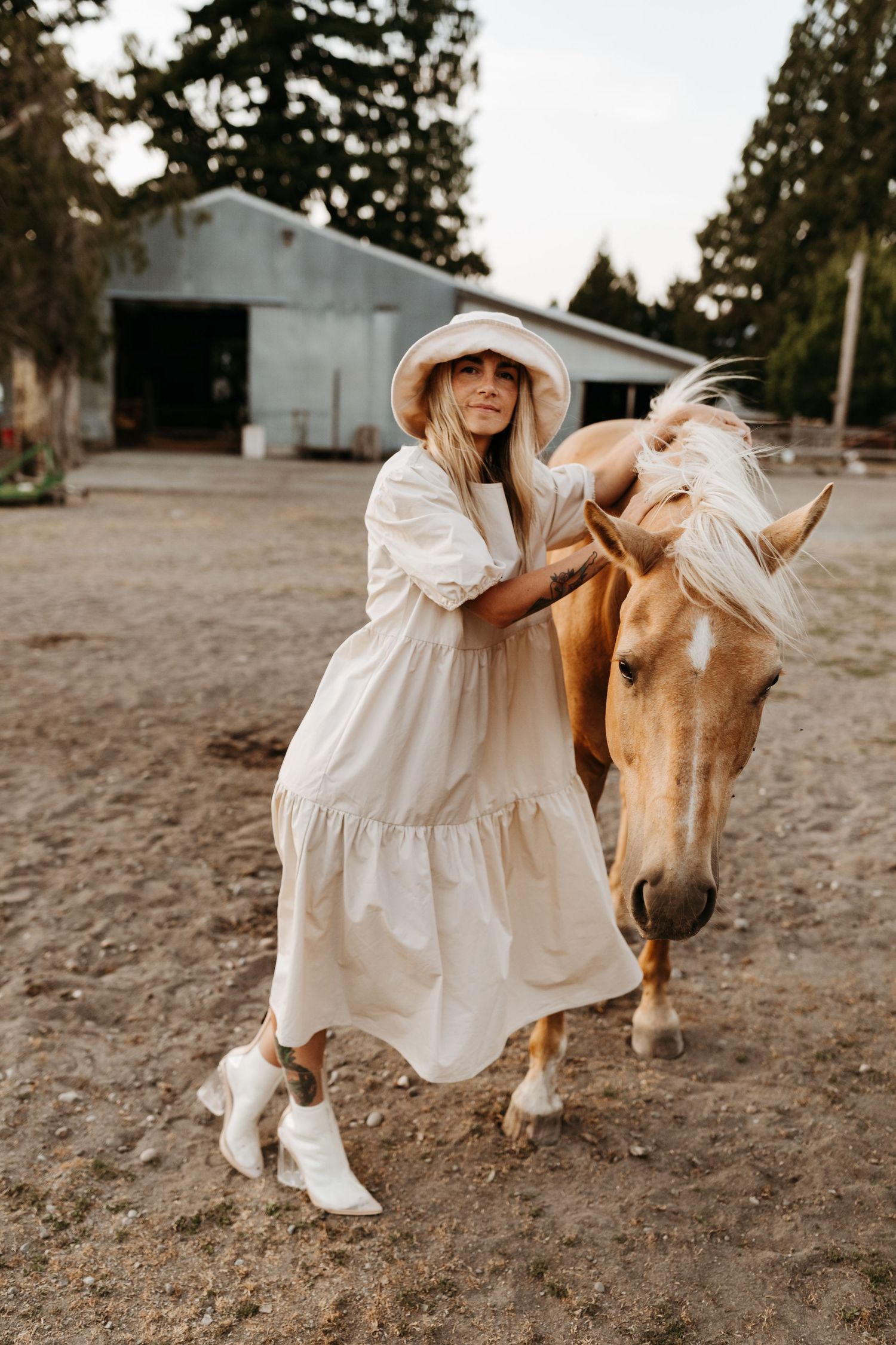 A person in a white prairie dress and sun hat poses with a palomino horse near a barn in rustic outdoor setting.
