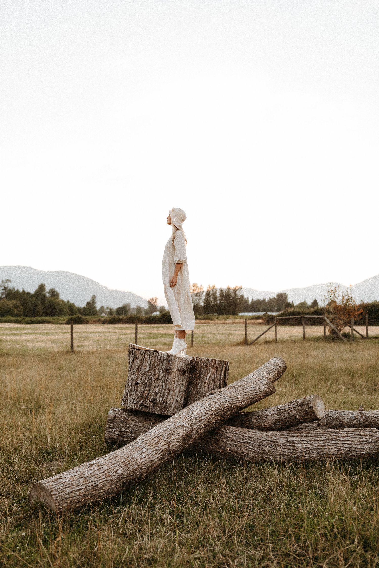 A figure in white standing on a large tree stump in a grassy field with mountains visible in the distance.