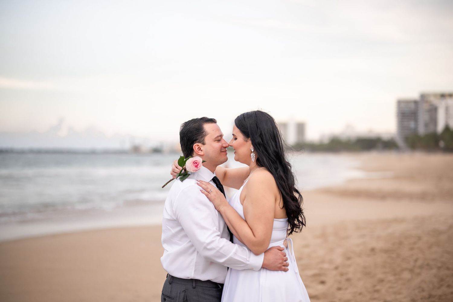 Romantic couple embrace on beach at sunset with ocean and cityscape in background.