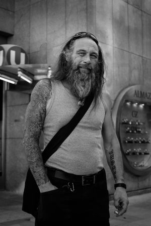 Black and white portrait of a bearded person wearing a tank top with visible tattoos on their arms.