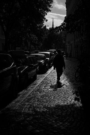 Silhouetted figure walking on cobblestone street with parked cars and church spire in background.
