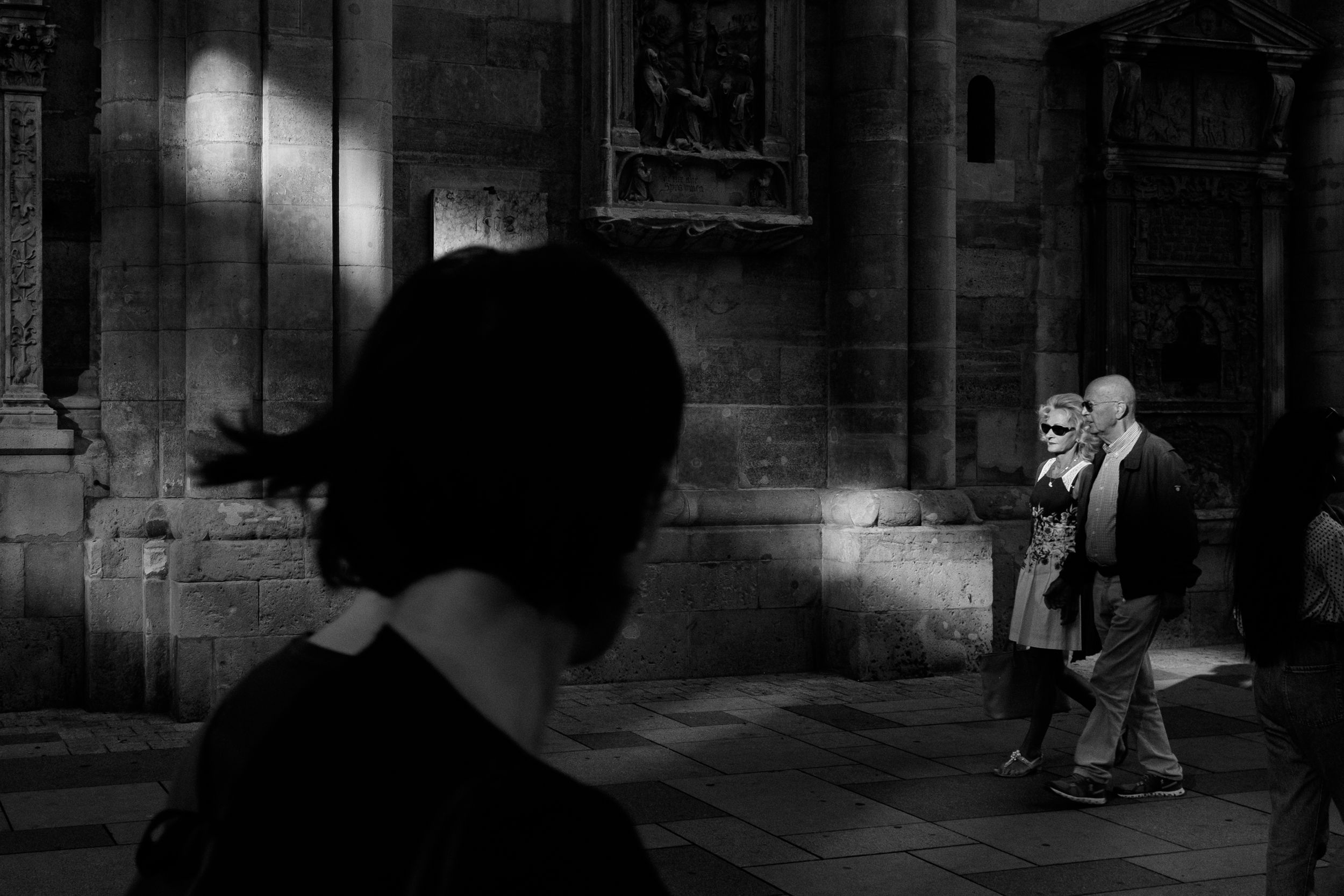 Film noir style black and white street photography with silhouettes casting dramatic shadows on a stone wall.