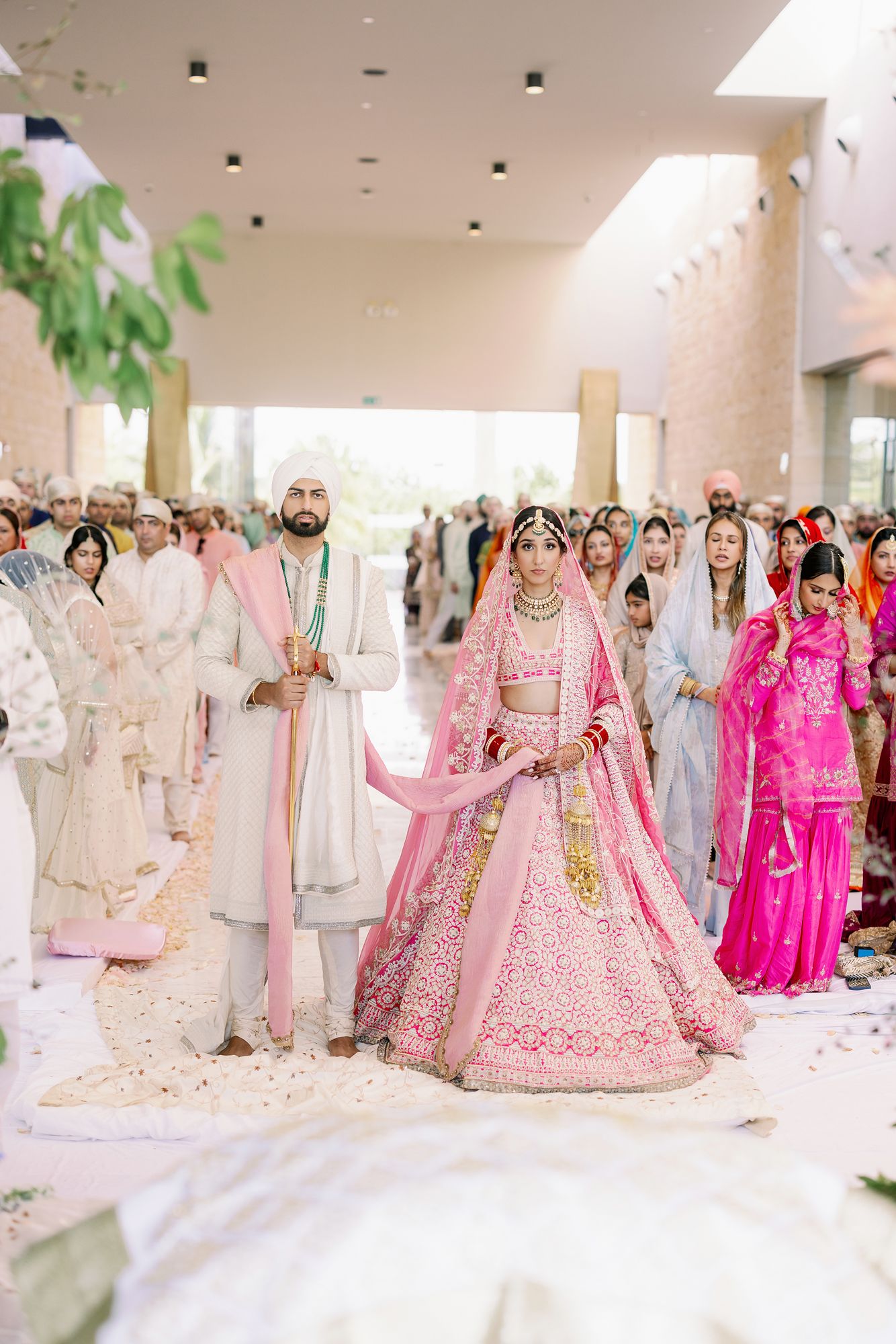Indian wedding ceremony with couple in pink and cream outfits surrounded by guests in white.