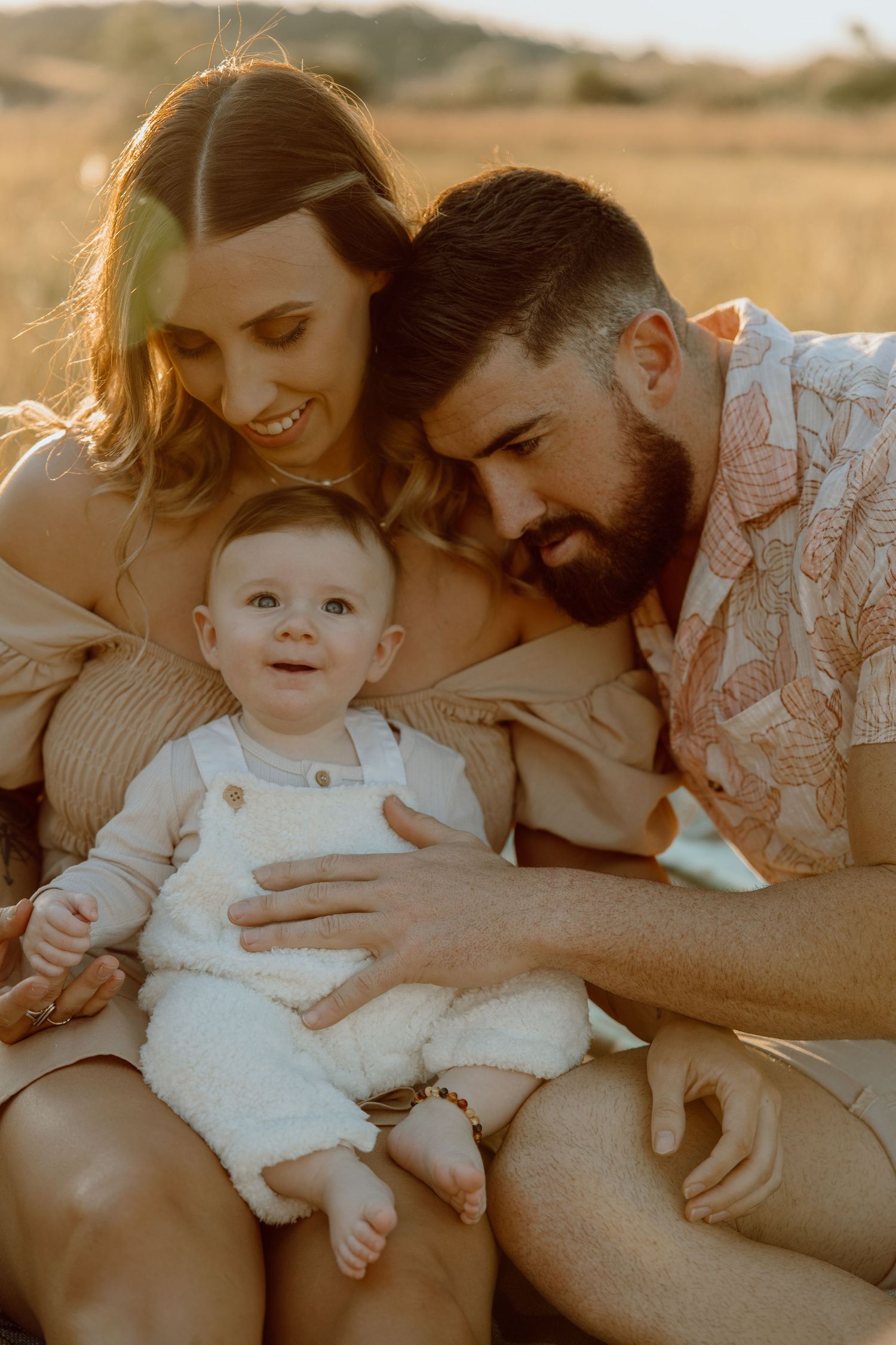Smiling mother and bearded father hold their happy baby in white overalls in warm golden light for a family portrait.