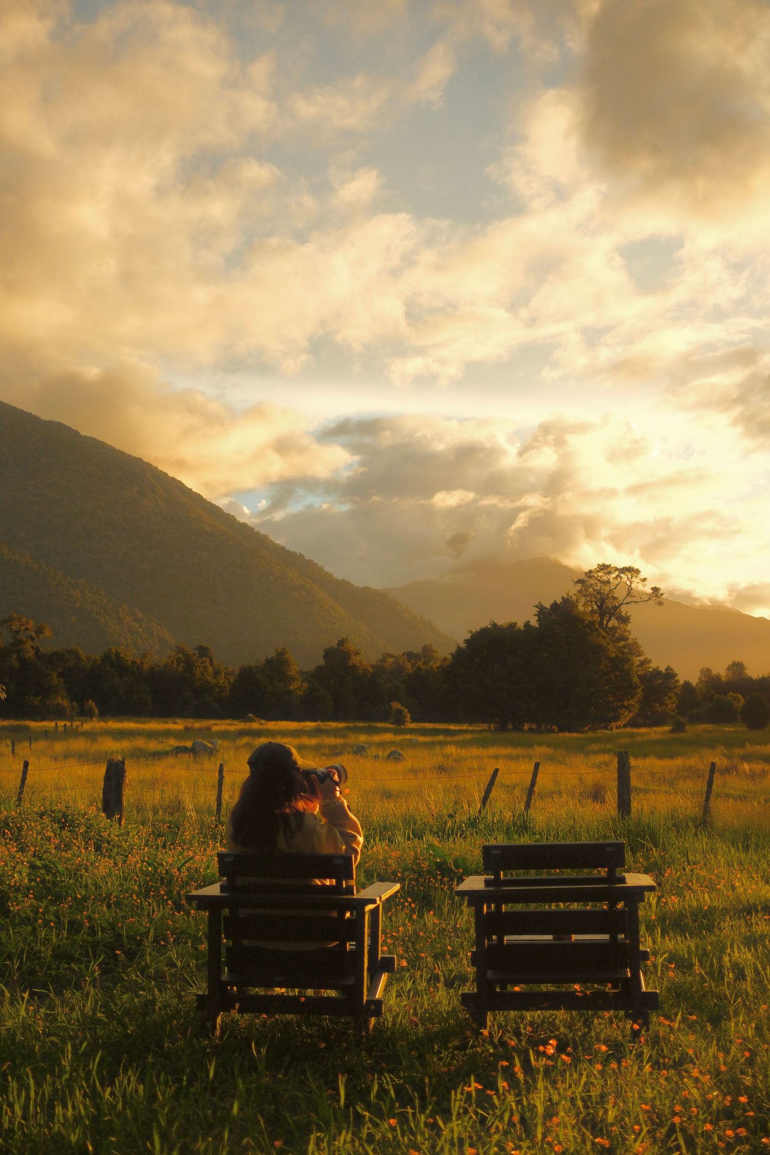 Person sits in a wooden chair holding a camera, facing a golden sunset over green pasture and misty mountains.